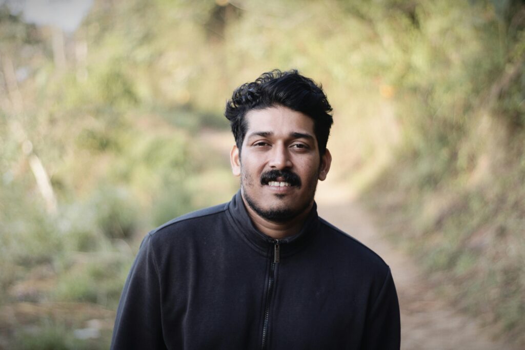 A portrait of a South Asian man in a black jacket standing outdoors on a natural pathway.